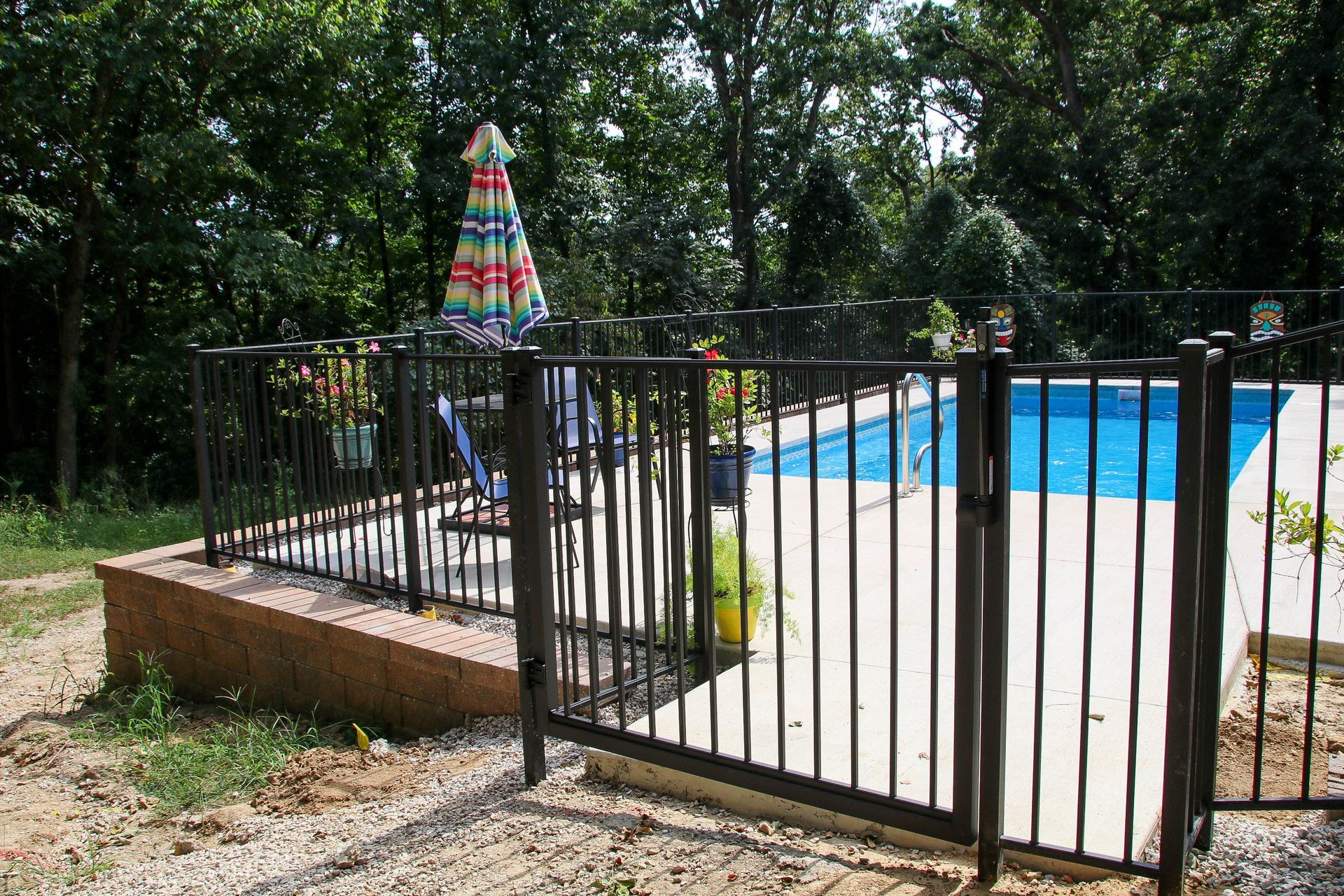 Black fence surrounds a swimming pool with an umbrella, chairs, and trees in the background.
