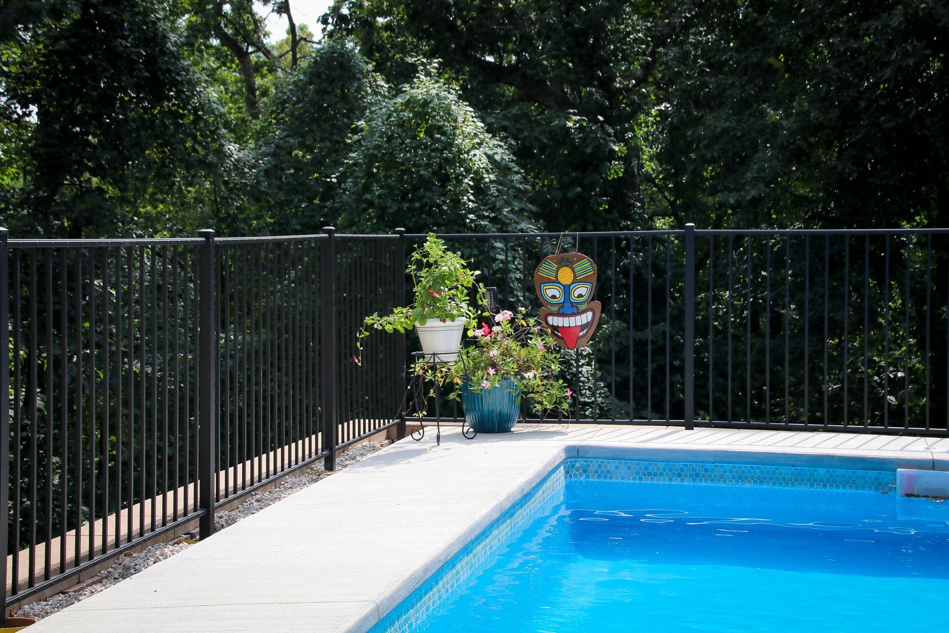 A pool with blue water next to a white deck, black fence, and trees. A plant and tiki mask sit on the deck.