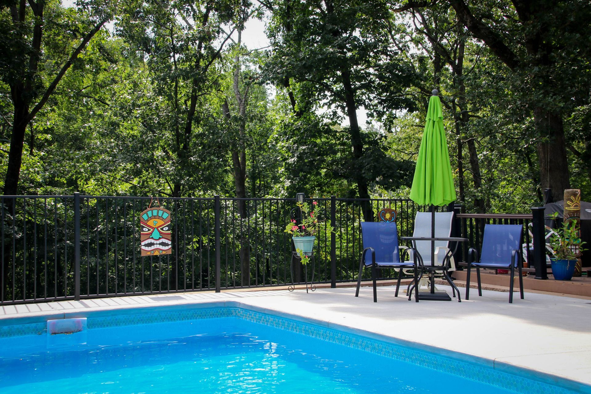 Poolside patio with table, chairs, and green umbrella, surrounded by trees.