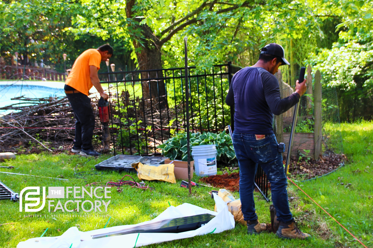 Two workers installing a black fence in a grassy yard near a tree. One uses a jackhammer.
