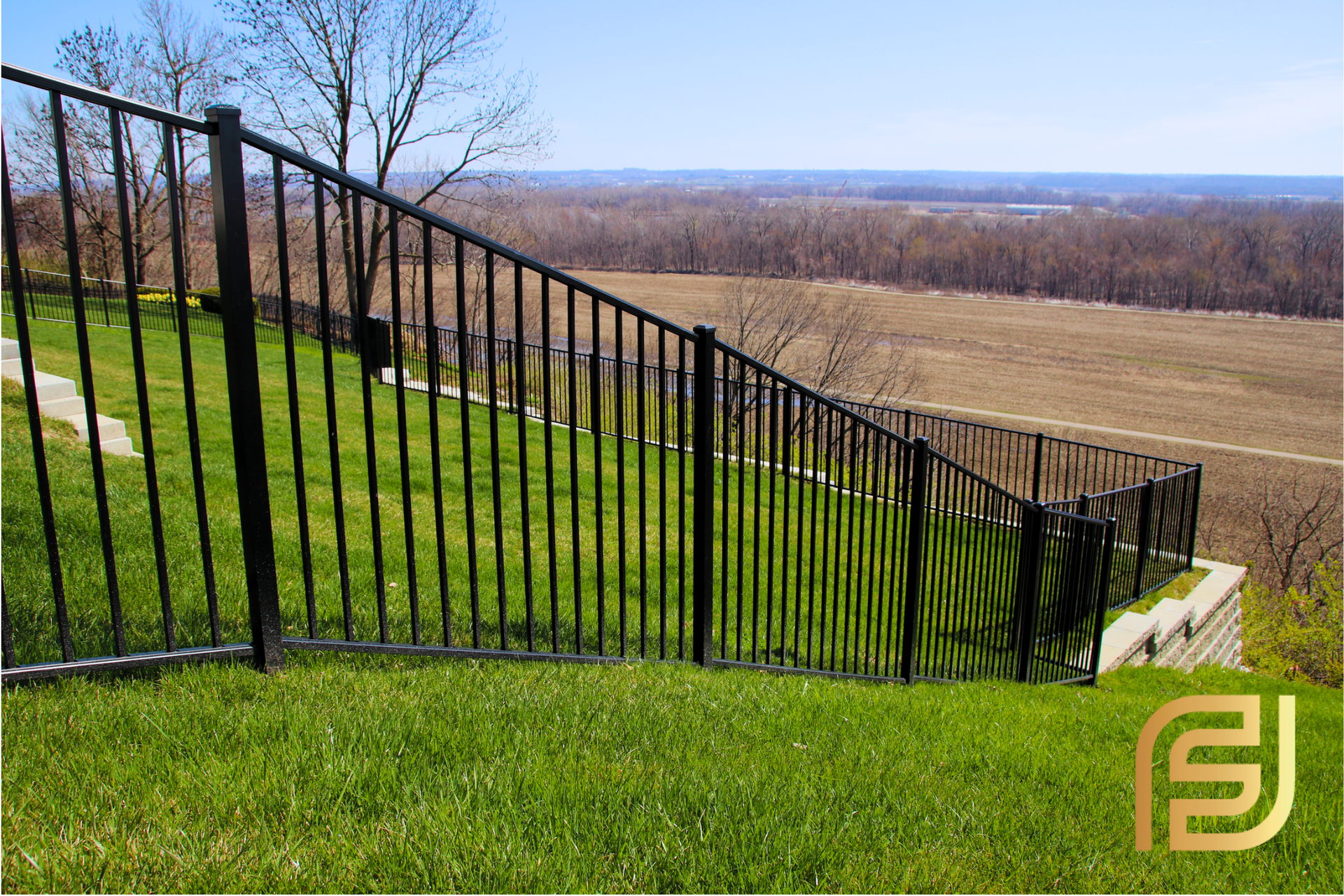 Black metal fence on a sloping green lawn, overlooking a distant rural landscape on a sunny day.
