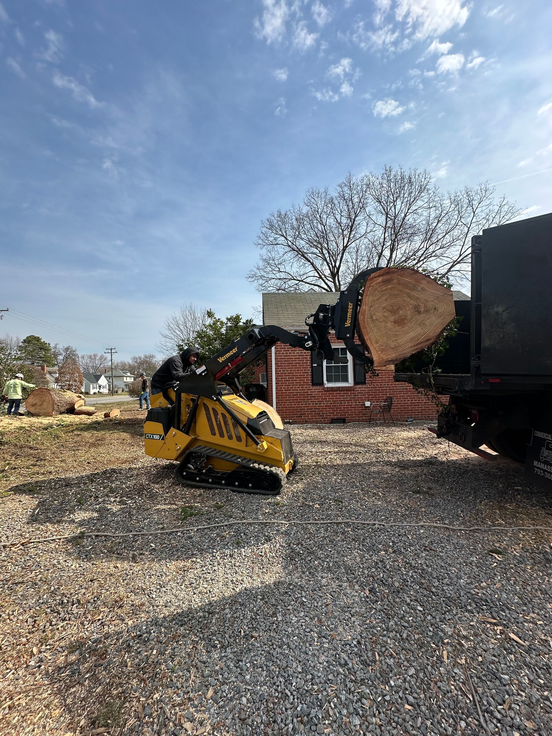 A yellow machine loads a large tree trunk into a truck; wood chips on the ground near a house under a blue sky.