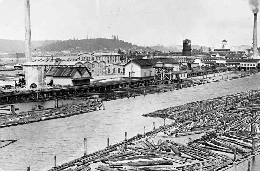 Industrial waterfront with logs floating in a river beside factory buildings and smokestacks.