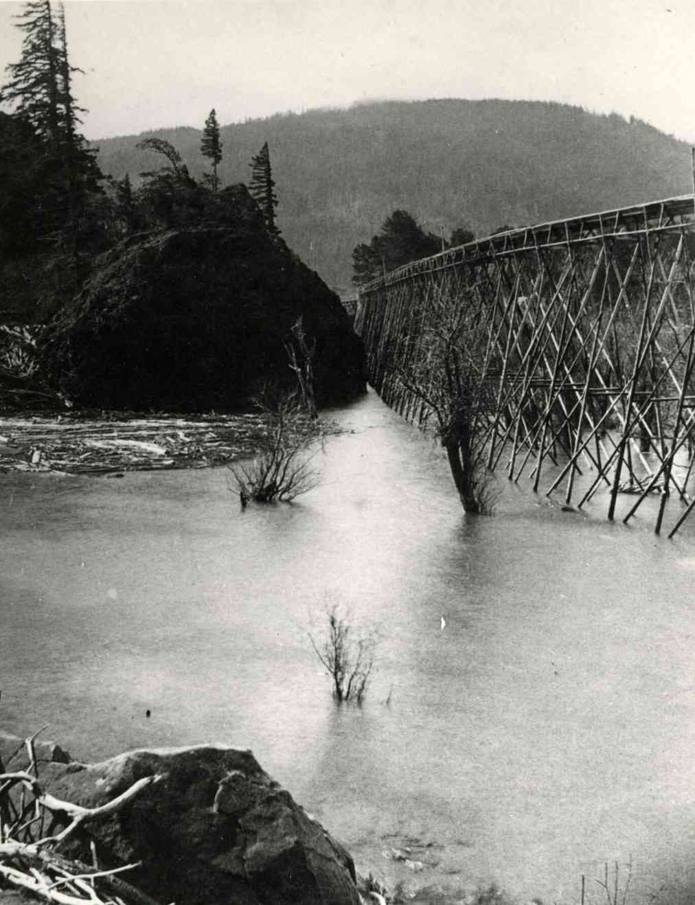 A river beside a wooden trestle bridge with mountains and trees in the background.
