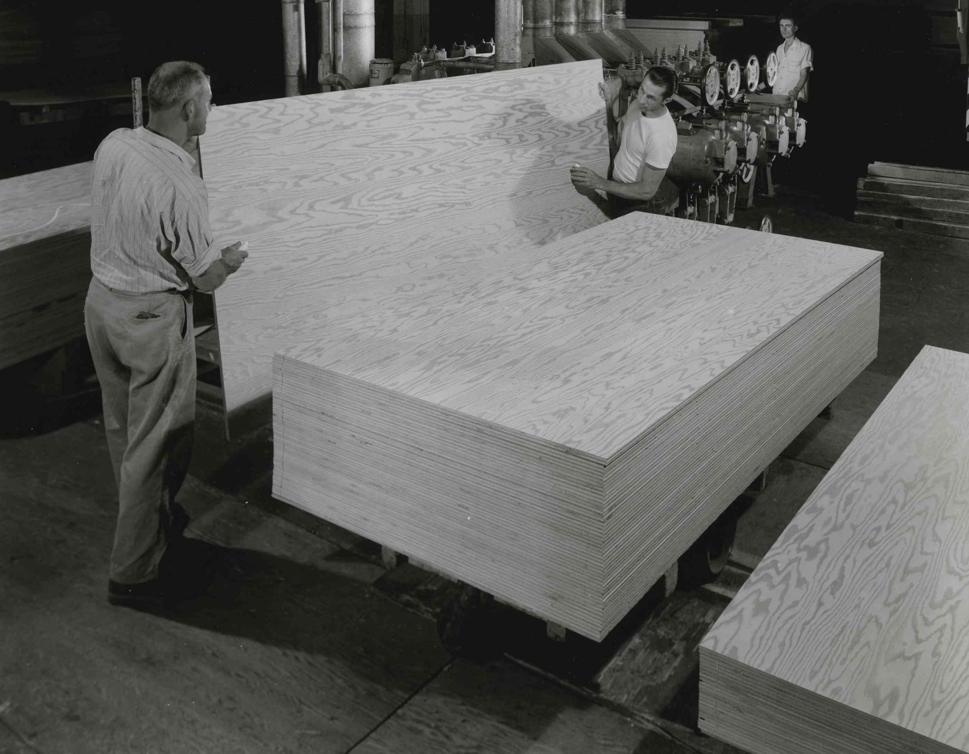 Workers moving large stacked plywood sheets in a factory or warehouse