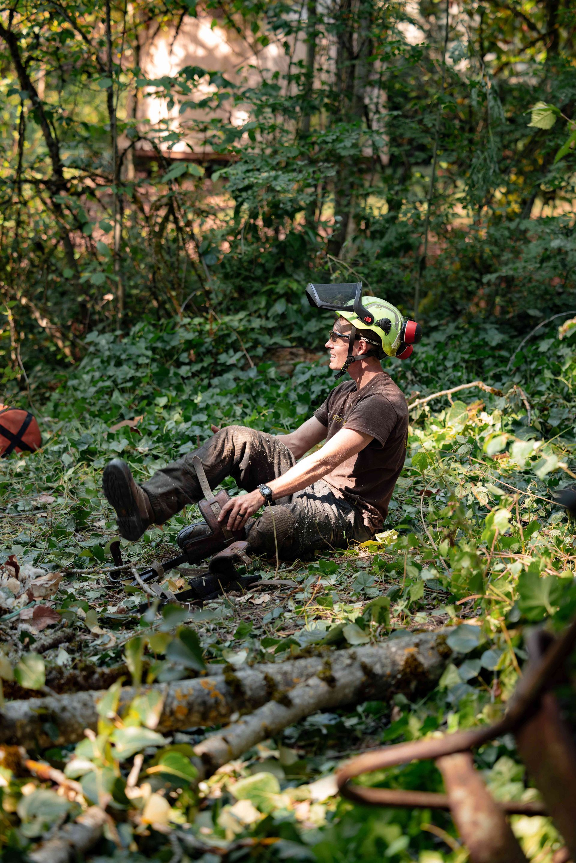 Person sitting on a leafy forest floor wearing a head-mounted camera, surrounded by greenery and trees.