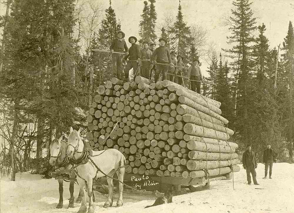 Horse-drawn log sled stacked with cut timber, workers standing on top in a snowy forest.