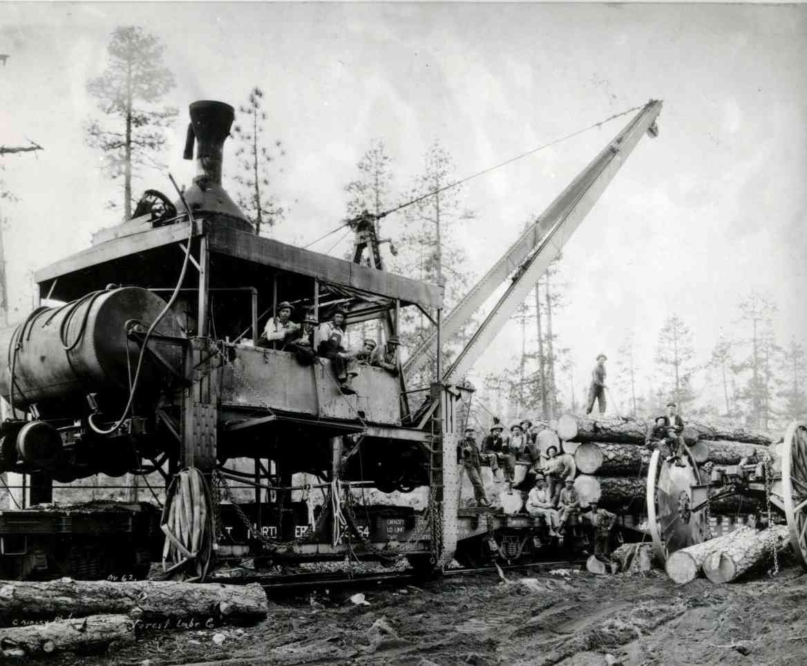 Black-and-white photo of a large logging machine with crane in a forest clearing, workers nearby and felled logs.