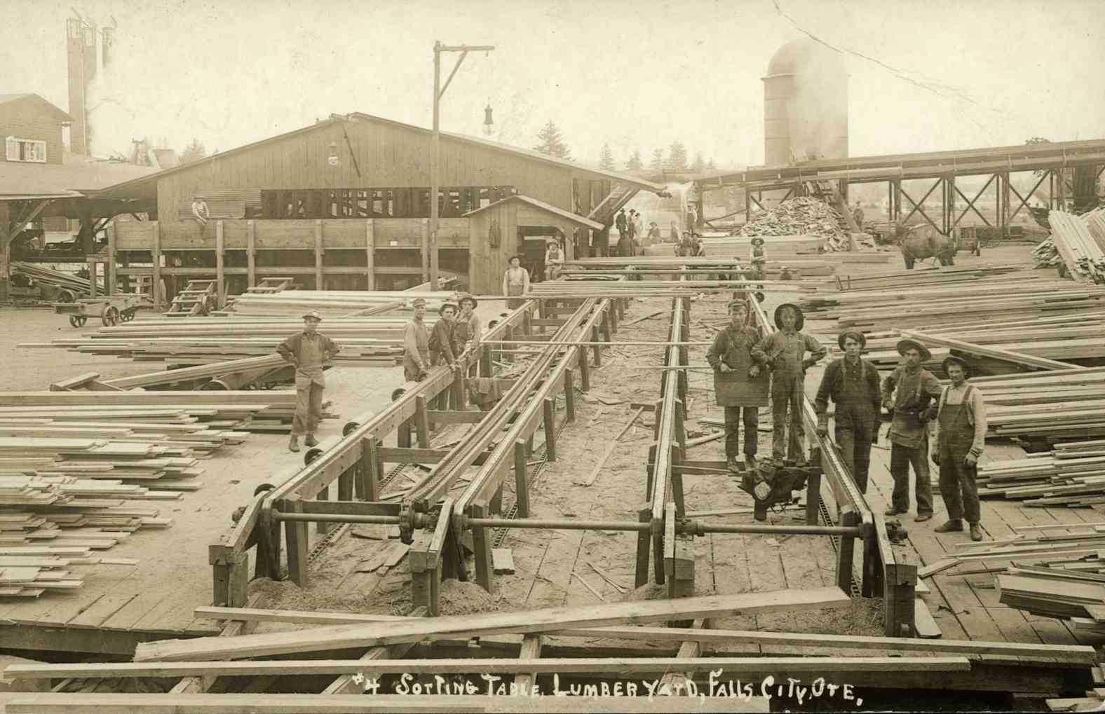 Historic lumber yard with workers stacking long wooden boards beside a mill and railway tracks.
