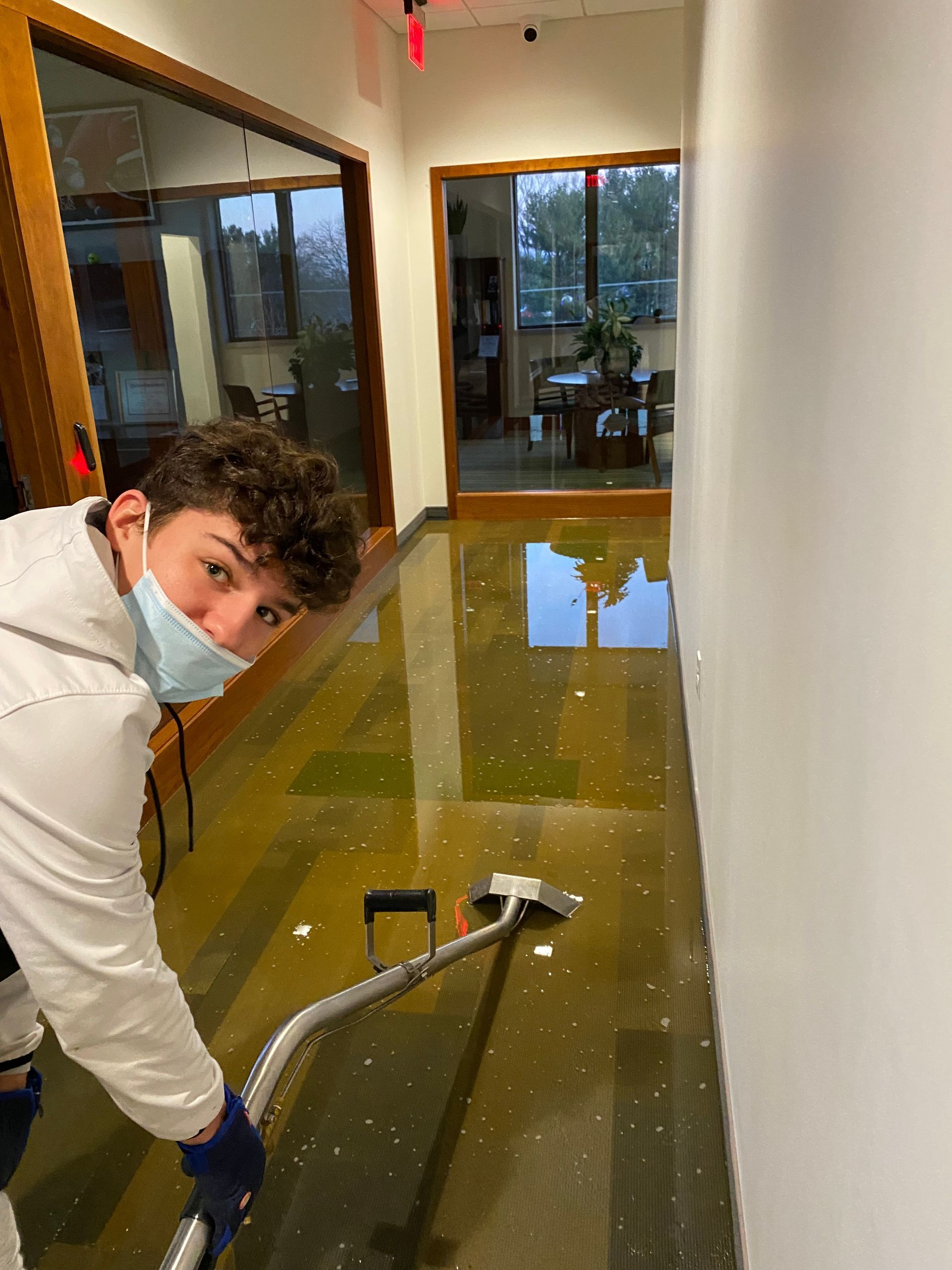 A man wearing a mask is cleaning a flooded floor with a vacuum cleaner.