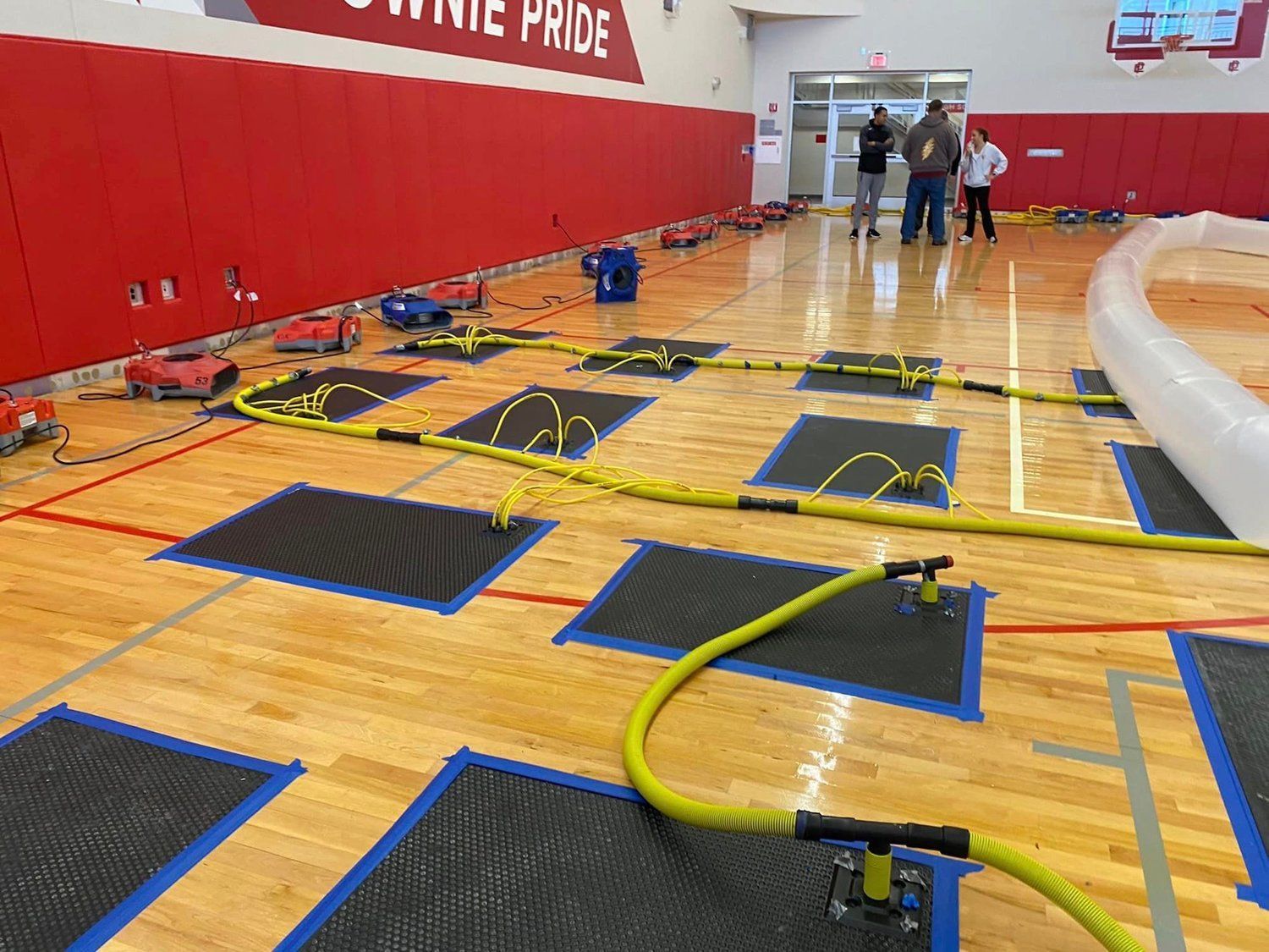 A group of people are standing on a wooden floor in a gym.