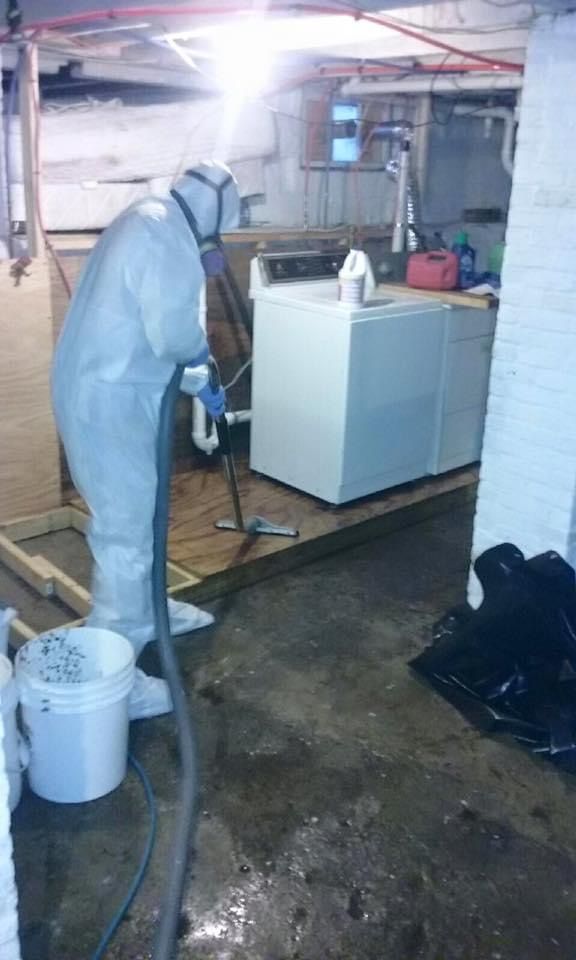 A man in a protective suit is cleaning a basement with a vacuum cleaner.