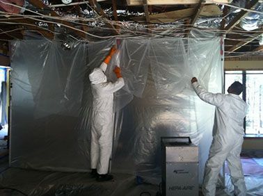 Two men in protective suits are covering a wall with plastic.