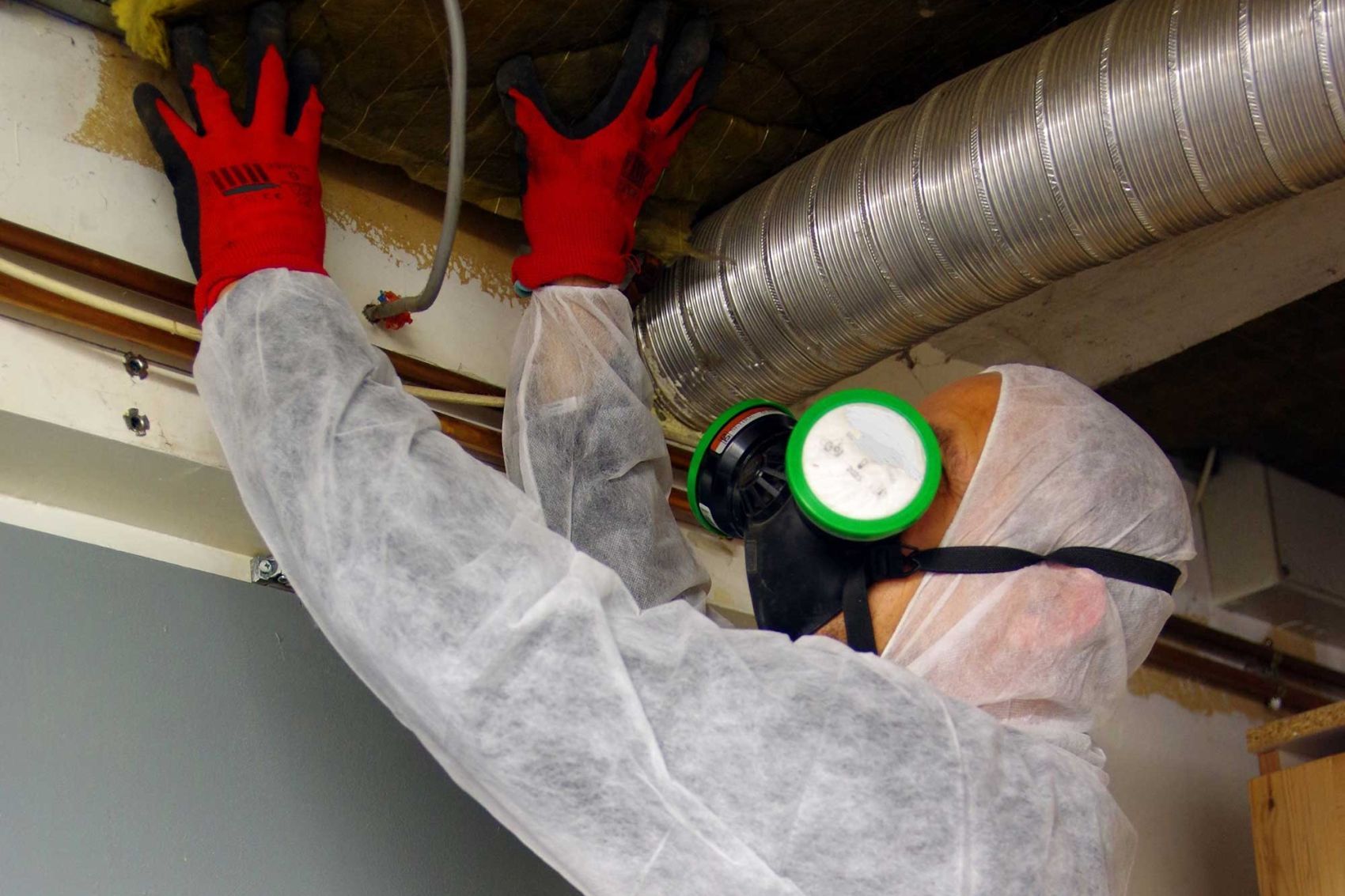 Person in protective gear working on ceiling, wearing mask and gloves.