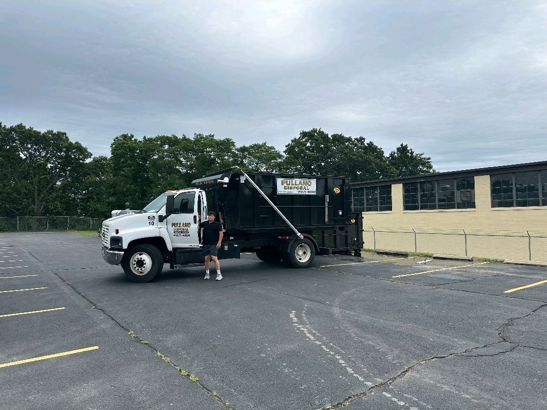 A man stands beside a black dump truck on a gray asphalt lot next to a building.
