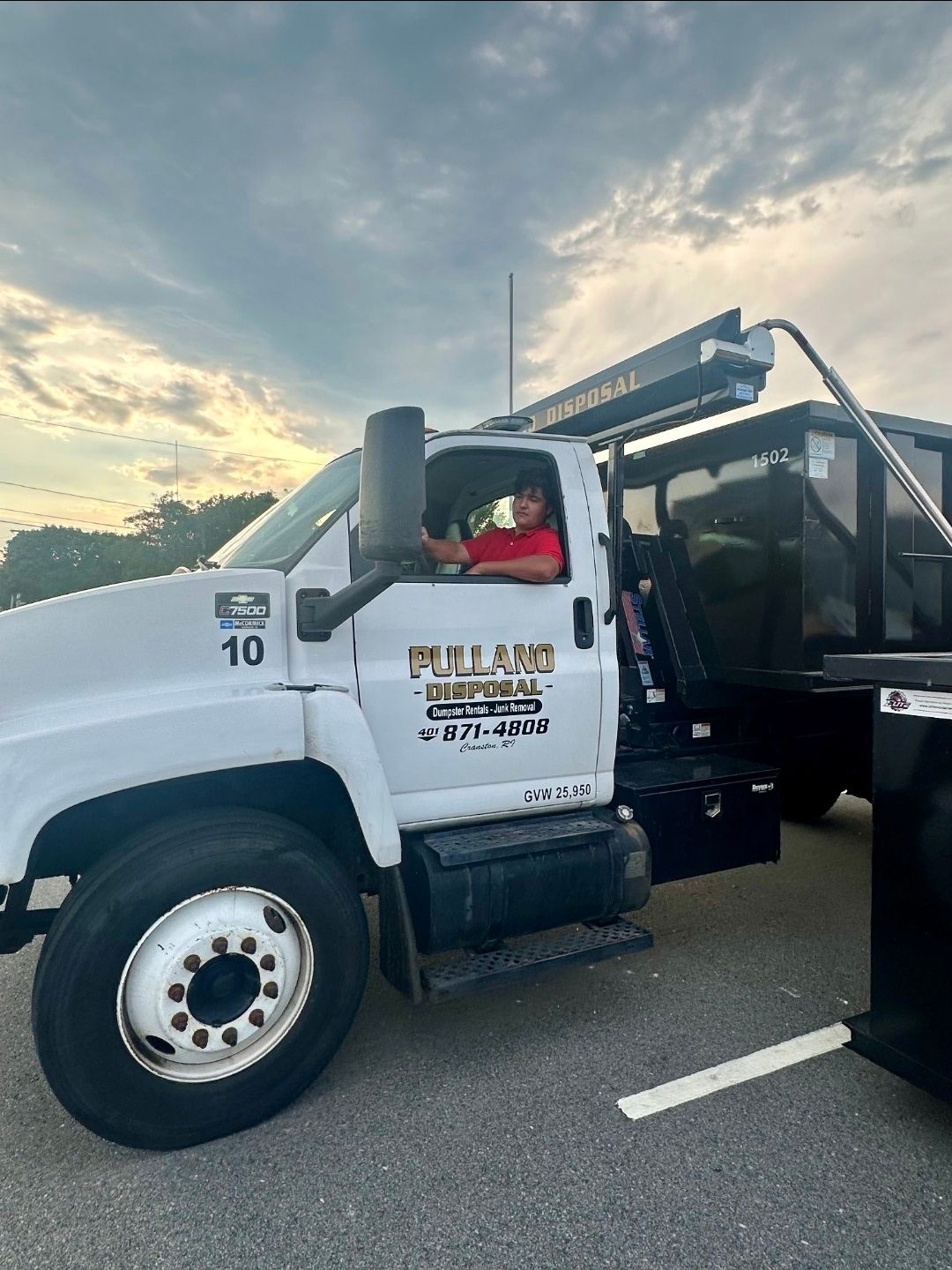 Man in red shirt in the cab of a white dump truck, outdoors. Truck has company logo. Cloudy sky.