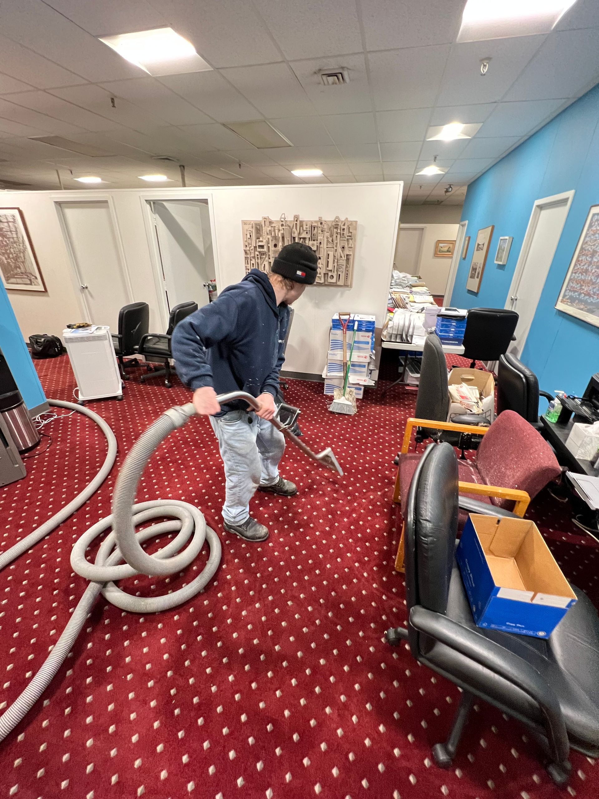 A man is using a vacuum cleaner to clean a red carpet in an office.