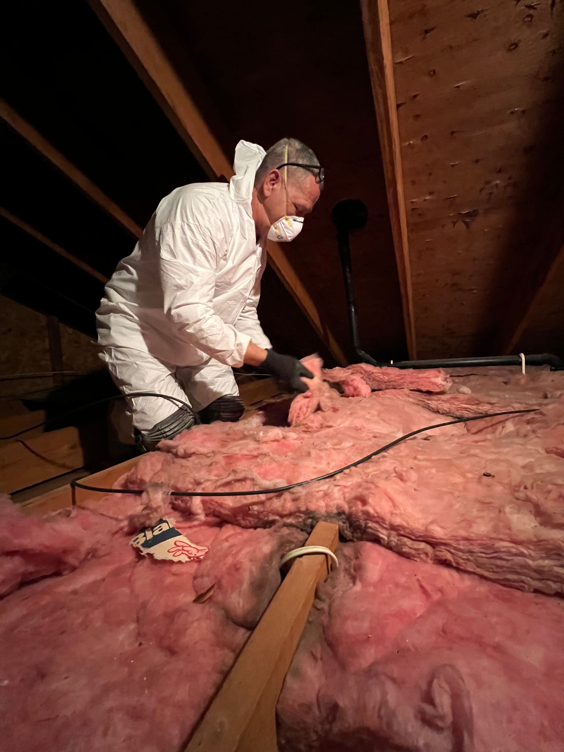A man wearing a mask is working on insulation in an attic.