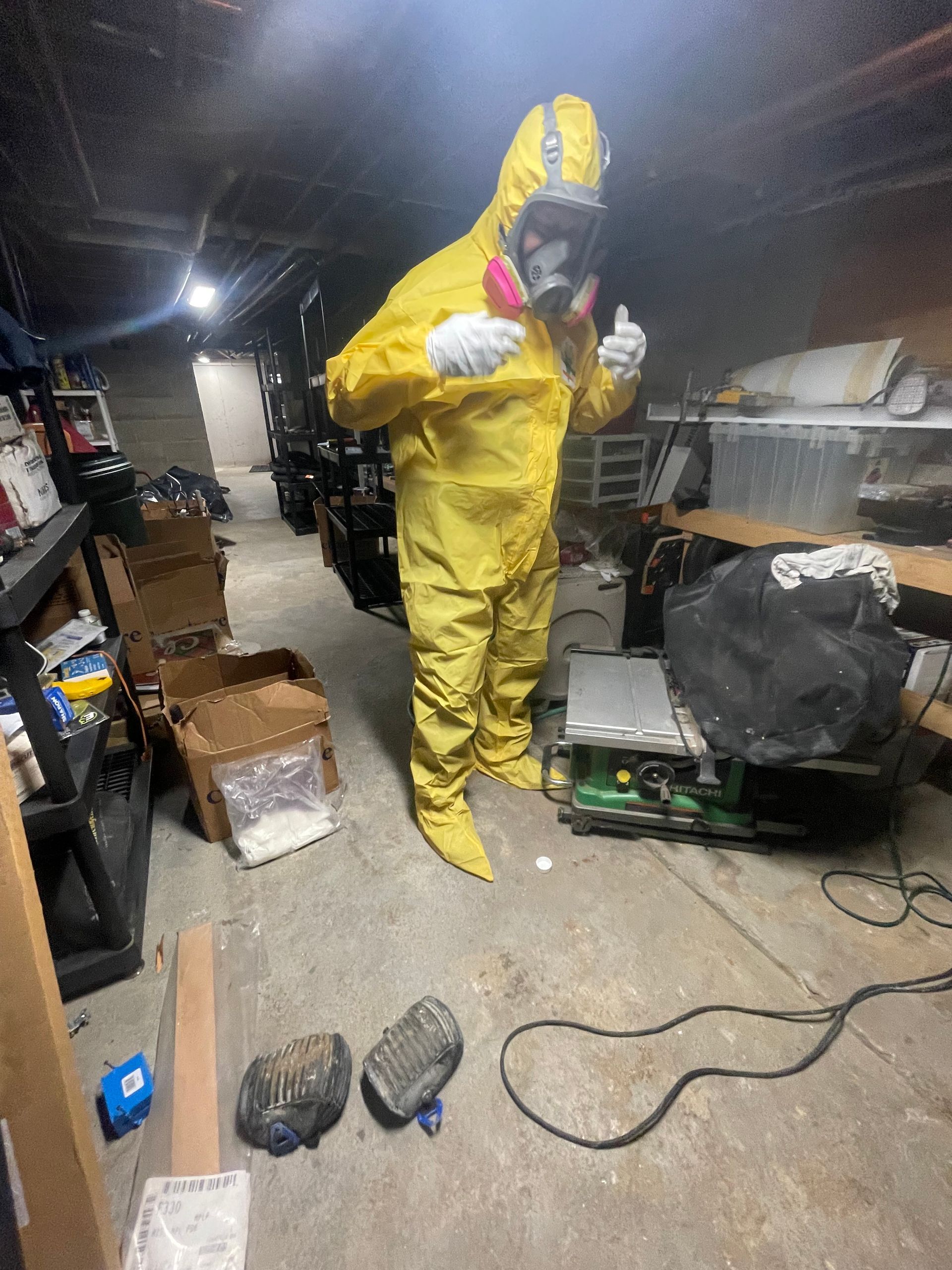 A man in a yellow suit and gas mask is standing in a messy room.