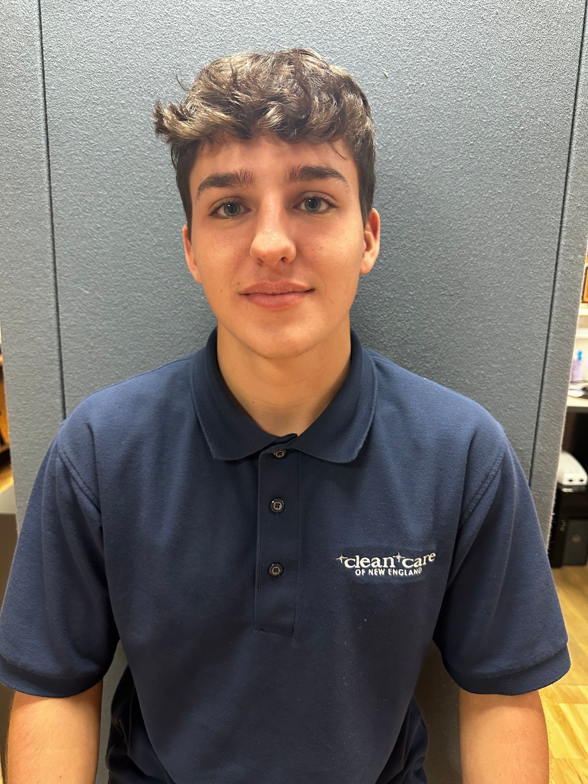 A young man wearing a blue polo shirt is sitting in front of a cubicle.