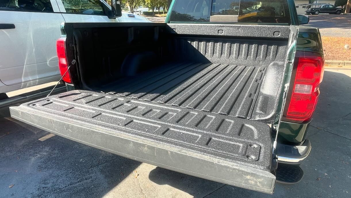 Open, black truck bed. Tailgate down, with protective liner. Green truck.