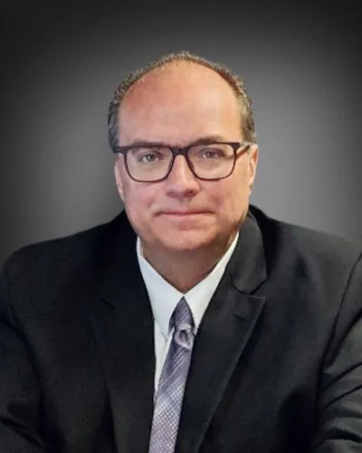 Man in suit and glasses, smiling, facing the camera against a gray gradient background.