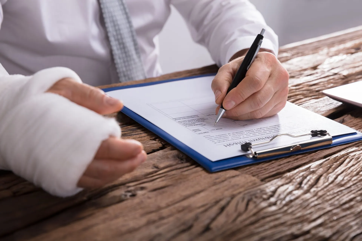 Person with a bandaged wrist writing on a clipboard at a wooden desk.