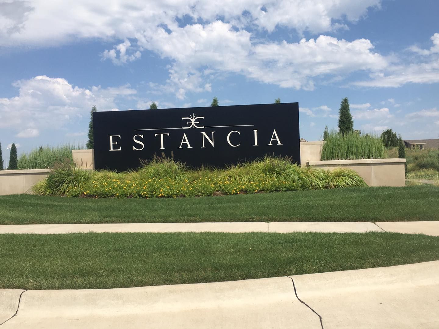 Sign for Estancia community with black background and white lettering, in front of green grass and blue sky.