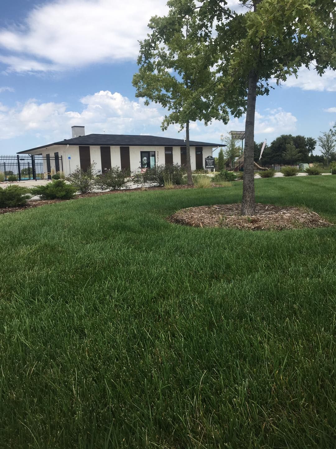 White building with black shutters and a dark roof, on green grass with a tree under a bright blue sky.
