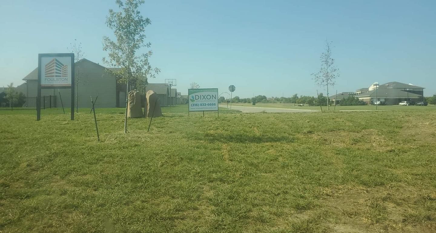 Grassy field with signs; houses in the background. Bright, sunny day.