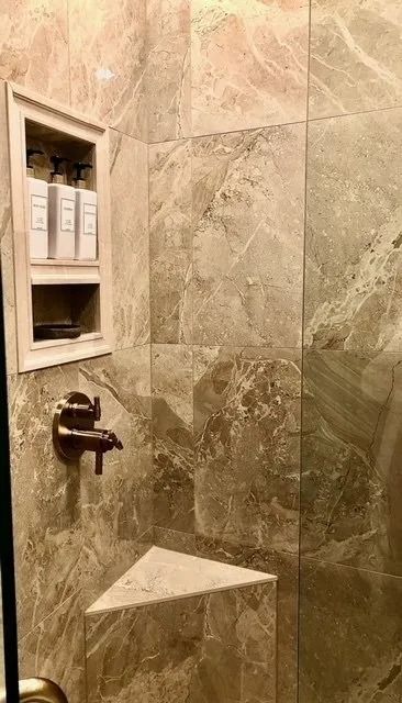 A tiled shower with a built-in soap niche and corner shelf. Bronze faucet and neutral-toned marble.