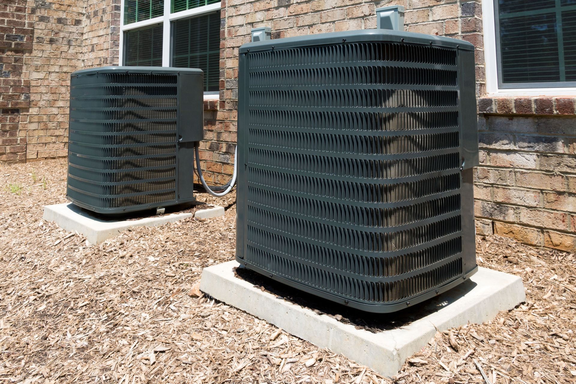 Two dark air conditioning units on concrete pads next to a brick building.