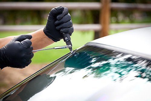 Hands in black gloves using tools to remove windshield seal from a car.