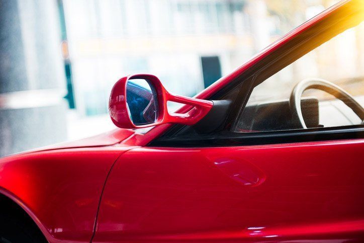 Red convertible car, driver's side detail: side mirror, steering wheel, open top. Bright sunlight.