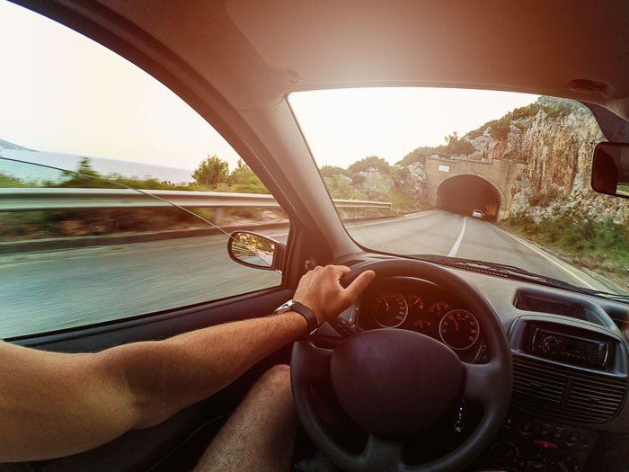 View from inside a car driving on a coastal road toward a tunnel. Driver's hand on the steering wheel.