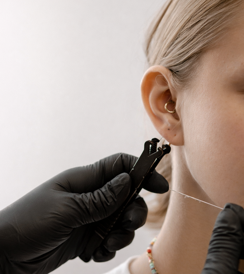 Person's ear being pierced, held with black gloved hands. Silver jewelry and tools are used in a sterile environment.