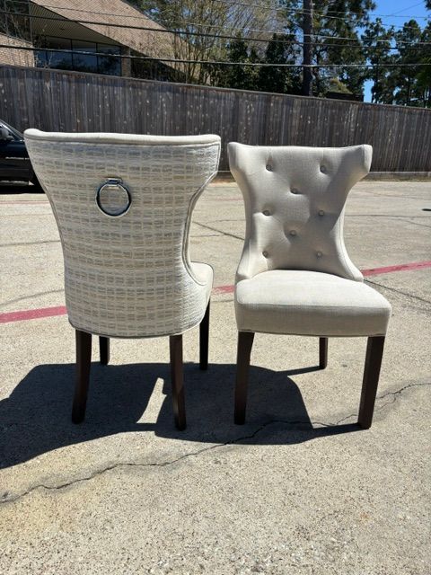 Two upholstered dining chairs outdoors on a sunlit concrete surface, one white woven chair and one tufted beige chair.