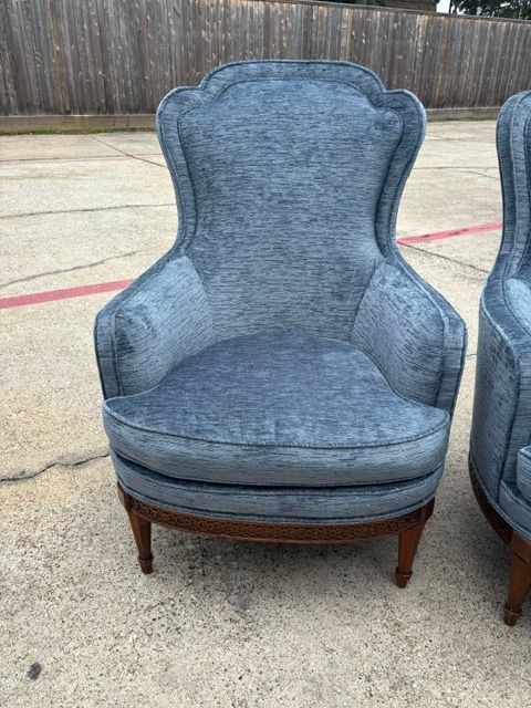 Blue upholstered wingback chair on a concrete surface outdoors, with another chair partly visible beside it.