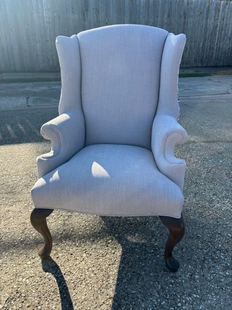 Light gray upholstered wingback chair with dark wooden legs sitting outdoors on pavement.