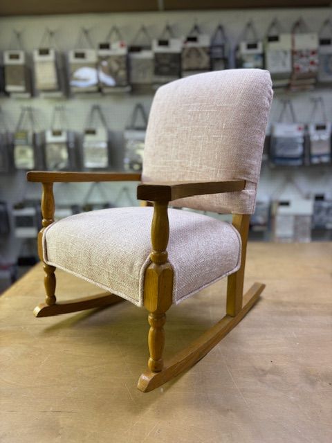 Child-sized light wood rocking chair with a beige upholstered seat and back. Background features fabric samples.