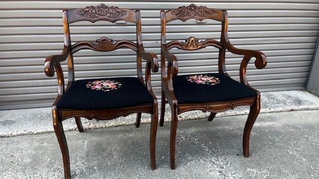 Two ornate wooden armchairs with black seats and floral embroidery against a gray backdrop.
