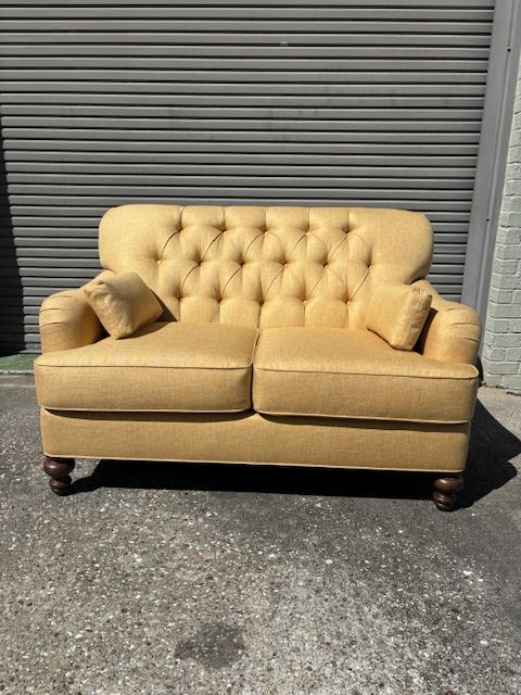 Yellow button-tufted loveseat with two matching pillows, on a concrete surface in front of a corrugated gray wall.