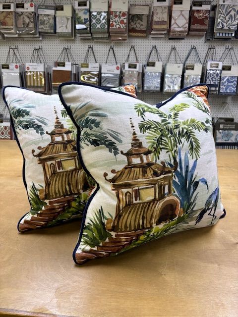 Two decorative pillows with Asian pagoda design, blue trim, on a wood surface, fabric samples in background.