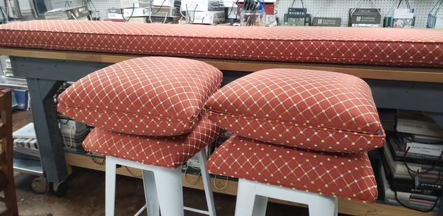 Red and white diamond patterned cushions stacked on white stools in a workshop, with a long cushion on a workbench behind them.