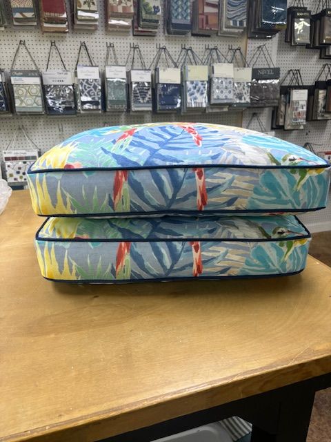 Two stacked tropical print cushions on a wooden table, surrounded by fabric samples in a store.