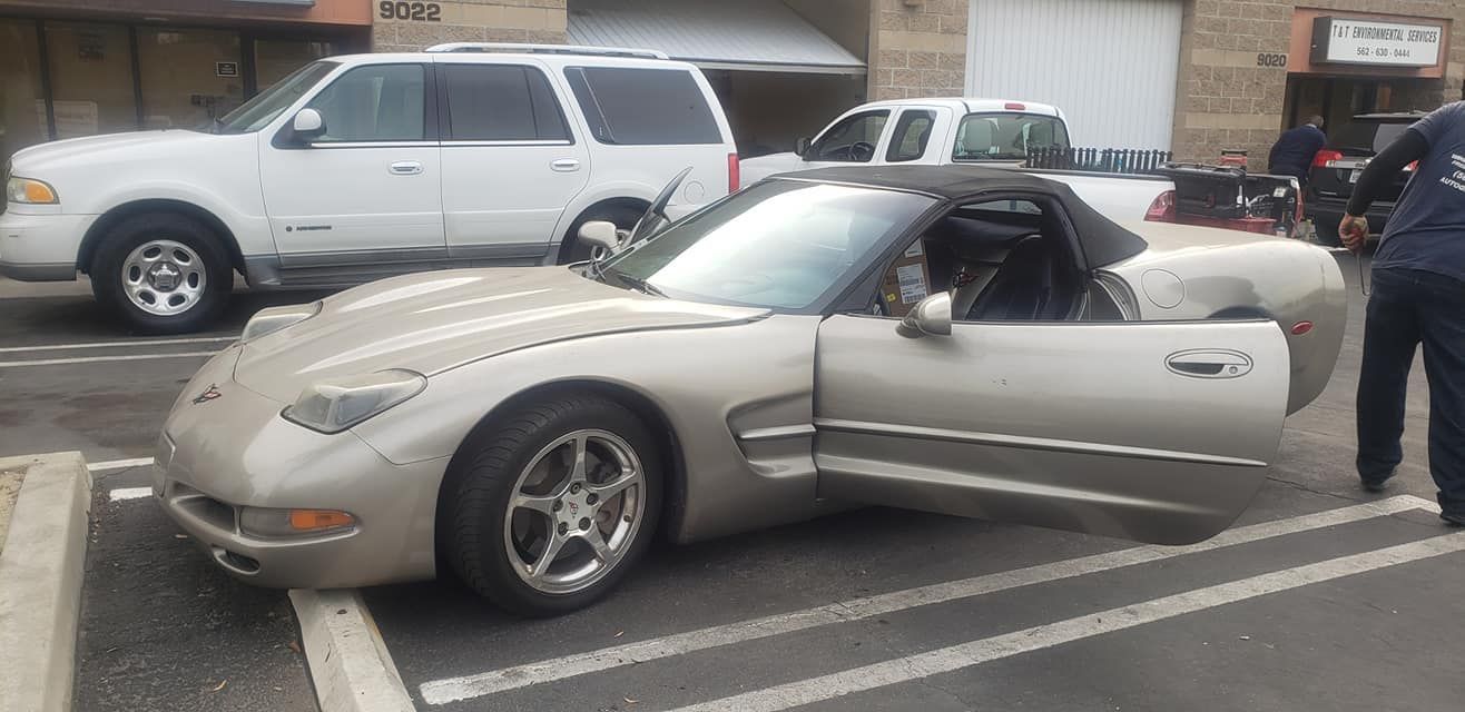 A silver convertible Corvette with an open door parked between white parking lines with a white SUV behind it.
