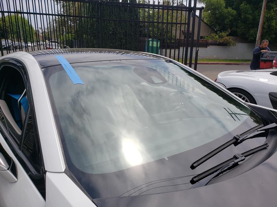 White car with black roof, blue tape on windshield. Sun reflecting in glass. Person standing nearby.