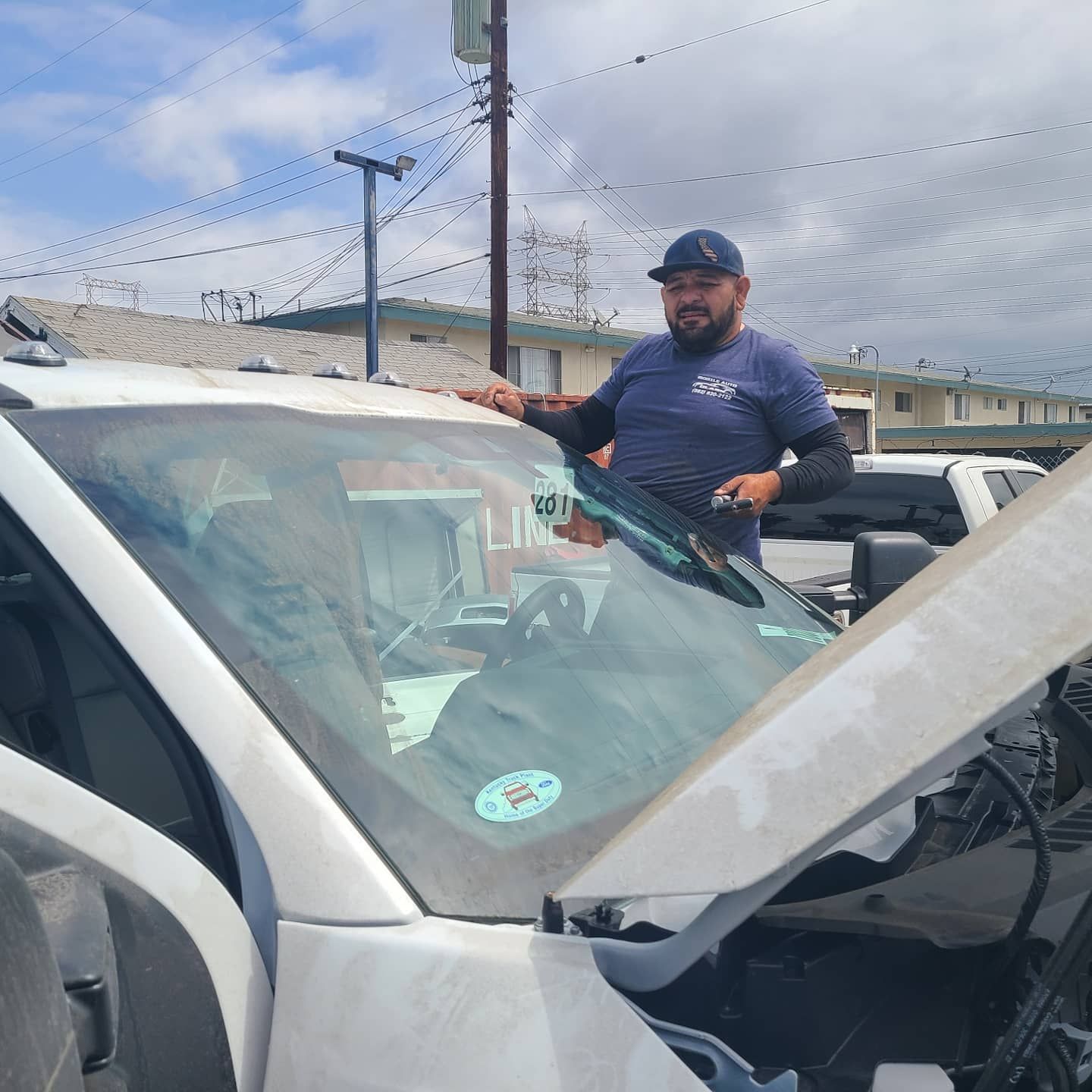 Man working on a car windshield, outdoors. He wears a hat and a shirt with a logo. Sunny day.