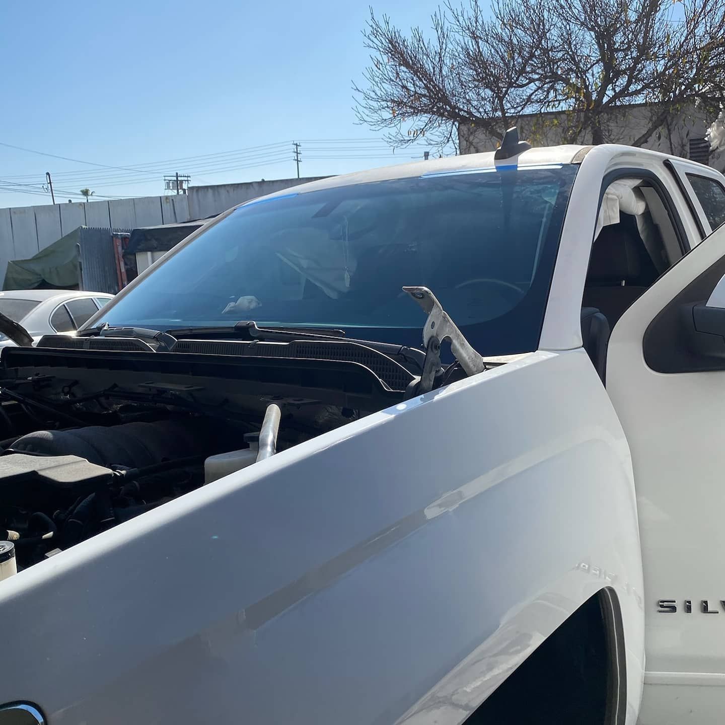 White truck with open hood and door at a salvage yard on a sunny day.