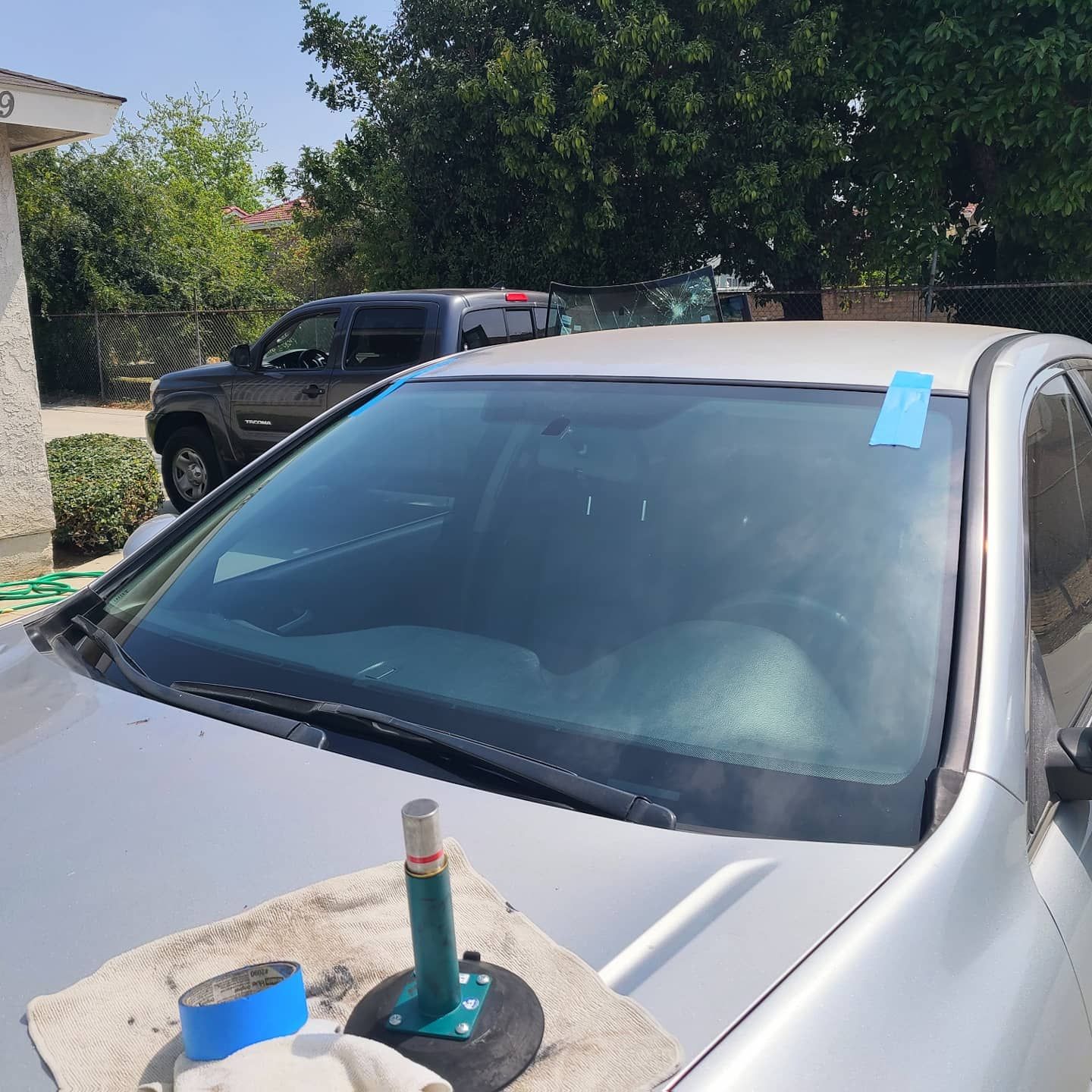 Silver car with blue tape on windshield; repair tools on hood, with trees and a truck in the background.